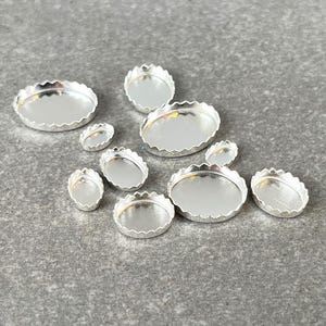 May include: A collection of small, silver-colored metal bezels in various oval and round shapes. Each bezel has a scalloped edge and a flat, reflective surface, perfect for jewelry making or crafting projects. The bezels are arranged on a gray surface.