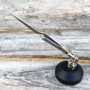 May include: A silver-toned tweezers tool with a black base. The tweezers are attached to a swiveling arm that allows for easy movement and adjustment. The base is weighted to provide stability.