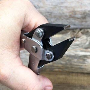 May include: A silver and black pliers tool with black rubberized jaws. The tool is held in a hand.