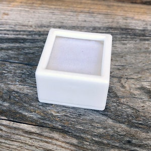 May include: A small, square, white jewelry box with a soft, white interior. The box is made of a smooth, solid material and has a simple, minimalist design. The box is empty and ready to hold a ring or other small piece of jewelry.