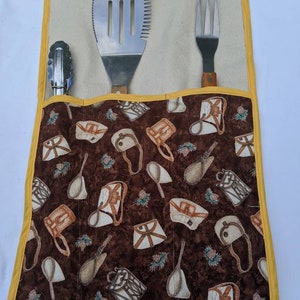 May include: A brown fabric grilling tool holder with a yellow trim. The holder has three pockets for tools. The tools include a spatula, tongs, and a fork. The fabric has a pattern of brown and white camping gear.