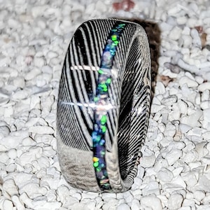 May include: A black and silver ring with a green and blue opal inlay. The ring is made of Damascus steel and has a unique, textured finish.
