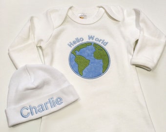 May include: White baby sleep gown and matching beanie. The gown features a blue and green embroidered globe design with the words "Hello World." The beanie is embroidered with the name "Charlie" in blue thread.