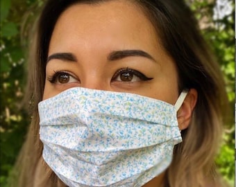 May include: A white face mask with a blue and green floral pattern. The mask has pleats and is made of fabric. The text "Blue Green Flower" is printed below the mask.