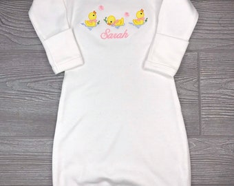 May include: White long-sleeved gown with pink trim and embroidered yellow ducks with pink bows. The gown has the name "Sarah" embroidered in pink below the ducks.