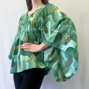May include: A green blouse featuring a geometric design and small orange floral details. The top has wide sleeves and a round neck. The fabric looks lightweight, with a flowing shape. The style is reminiscent of vintage fashion.