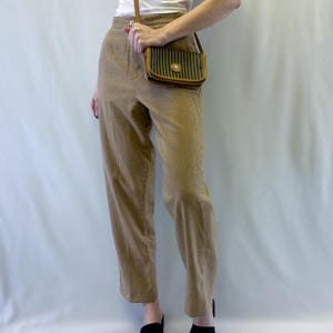 May include: A pair of tan high-waisted trousers, a white top, and black sandals. A small, brown and green striped crossbody bag with a gold clasp is worn over the shoulder. The outfit is simple and classic.