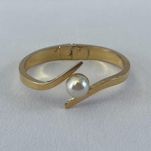 May include: A gold-colored bangle bracelet with a white pearl accent. The bracelet has a hinged clasp and a curved design that cradles the pearl. The pearl is a round, off-white color.
