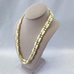 May include: A cream-coloured necklace with a double strand design. The necklace features elongated, rounded beads separated by smaller, round beads. The necklace is displayed on a mannequin bust.