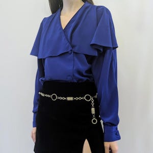 May include: A royal blue blouse with a large, dramatic collar and long sleeves. The blouse has a button closure at the front and is made of a silky fabric. The model is wearing a gold chain belt with rhinestone accents.