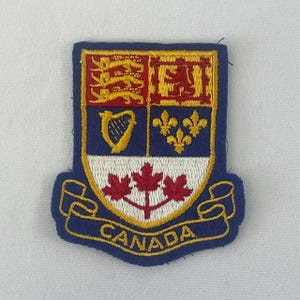 May include: An embroidered patch featuring a shield divided into four quadrants with three gold lions, a red lion, a gold harp, and three gold fleurs-de-lis. Below, three red maple leaves sit on a white field, with a banner reading CANADA.