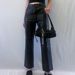 May include: Black leather trousers with a high waist and straight leg cut, paired with a black nylon shoulder bag featuring a prominent white tag with the text BAGGU printed in black capital letters hanging from the silver metal hardware.