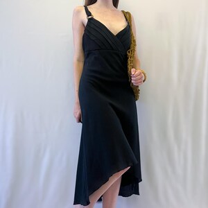 May include: A black midi dress featuring a V-neckline with pleated bodice detailing and thin shoulder straps connected by circular rings. The skirt has an asymmetrical hemline, and the garment is paired with a textured mustard yellow shoulder bag.