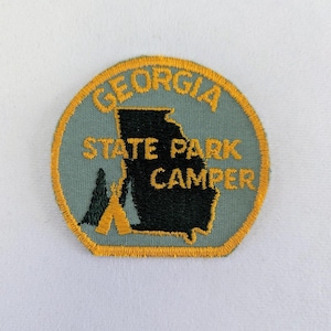 May include: A round, blue and yellow embroidered patch with the text "Georgia State Park Camper" and an outline of the state of Georgia. Two yellow tents are in the bottom left corner.