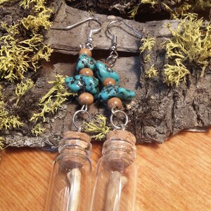 Op de afbeelding: Een paar oorbellen met turquoise en bruine kralen die hangen aan kleine glazen flesjes met een bot erin. De flesjes zijn voorzien van kurken.