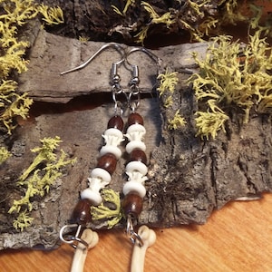 Op de afbeelding: Een paar oorbellen met bedels in de vorm van botten en bruine en witte kralen. De oorbellen hangen aan zilveren haakjes.