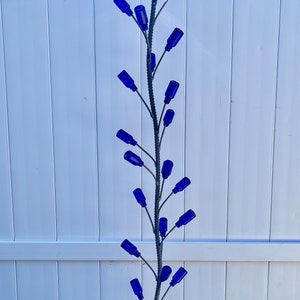 May include: A tall, vertical metal garden stake featuring a central rebar pole with numerous thin, branching metal arms. Each arm holds a small, vibrant cobalt blue glass bottle at its tip, creating a tree-like structure with blue glass accents.