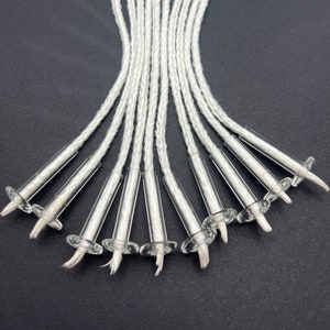 May include: A close-up of ten white braided candle wicks with clear glass bases. The wicks are arranged in a fan shape.