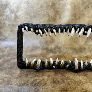 May include: Black metal license plate frame with a row of silver teeth along the top and bottom edges.
