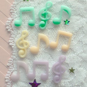 May include: A collection of colorful plastic music notes and treble clefs on a white lace background with iridescent glitter stars.