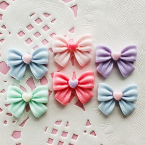 May include: A collection of colorful resin bows in pastel shades of blue, pink, green, and purple, each with a heart-shaped center. The bows are arranged on a white lace doily against a pink background.