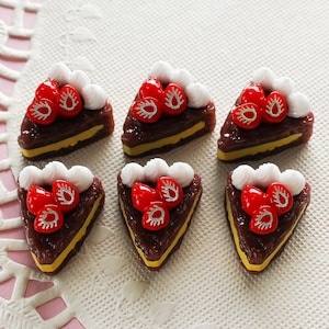 May include: Six miniature strawberry cake slices. Each slice features a dark brown cake base with a yellow layer, topped with white frosting and red strawberries. The cakes are arranged on a white, textured surface.