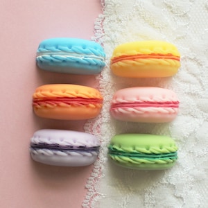 May include: Six colorful macarons arranged in two rows of three on a pink and white background. The macarons are in shades of blue, orange, pink, purple, and green.