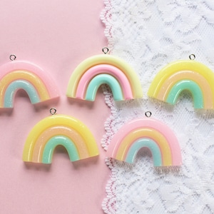 May include: Five pastel rainbow charms with silver jump rings. The rainbows are made of resin and have a layered effect with yellow, pink, and green colors.