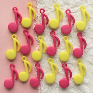 May include: A collection of pink and yellow plastic music note charms. The charms are arranged in a grid pattern on a white lace background.