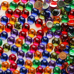 May include: A large assortment of colorful round glass gems in various shades of red, orange, yellow, green, blue, purple, and clear. The gems are scattered on a white doily.
