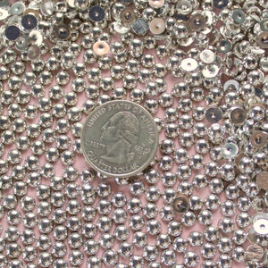 May include: A close-up image of a large quantity of silver-colored round beads. The beads are arranged in a random pattern and are all the same size. A US quarter is included in the image for scale.