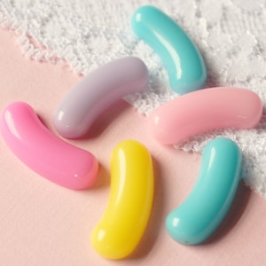 May include: Six jelly bean shaped candies in various colors: light blue, yellow, pink, light purple, and two shades of pink. The candies are arranged on a pink surface.