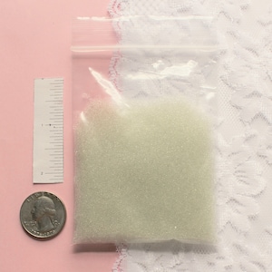 Op de afbeelding: Een klein doorzichtig plastic zakje gevuld met witte glazen kralen. Het zakje ligt op een roze oppervlak naast een liniaal en een Amerikaanse kwart.
