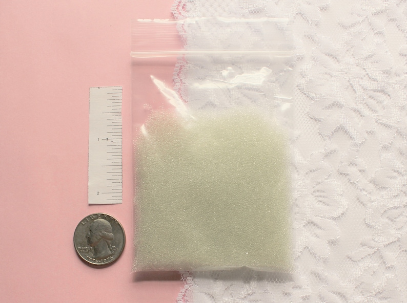 Op de afbeelding: Een klein doorzichtig plastic zakje gevuld met witte glazen kralen. Het zakje ligt op een roze oppervlak naast een liniaal en een Amerikaanse kwart.