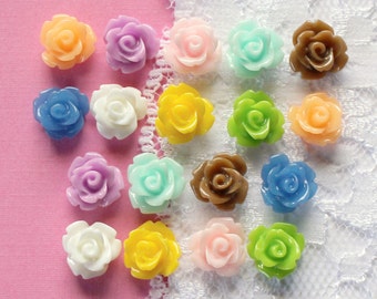 May include: A collection of small, colorful rose-shaped embellishments in various shades, including orange, purple, pink, blue, white, yellow, green, and brown. These decorative elements are arranged on a pink surface with a white lace trim.