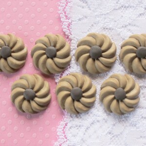 May include: Seven beige, flower-shaped embellishments with a gray center are arranged on a pink polka-dot background with a white lace trim. The embellishments have a spiral design.