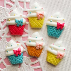 May include: A collection of colorful, miniature ice cream cone decorations. Each cone features a white ice cream swirl, a bow, and a waffle cone base in various colors, including red, yellow, orange, and blue. Small, colorful dots are scattered on the ice cream.