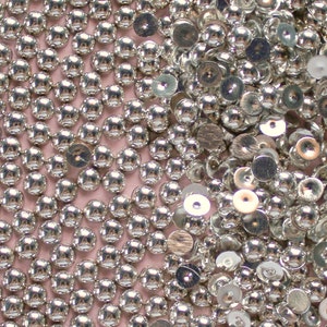 May include: A close-up image of a large quantity of silver-colored round and flat sequins. The sequins are scattered randomly on a pink surface.