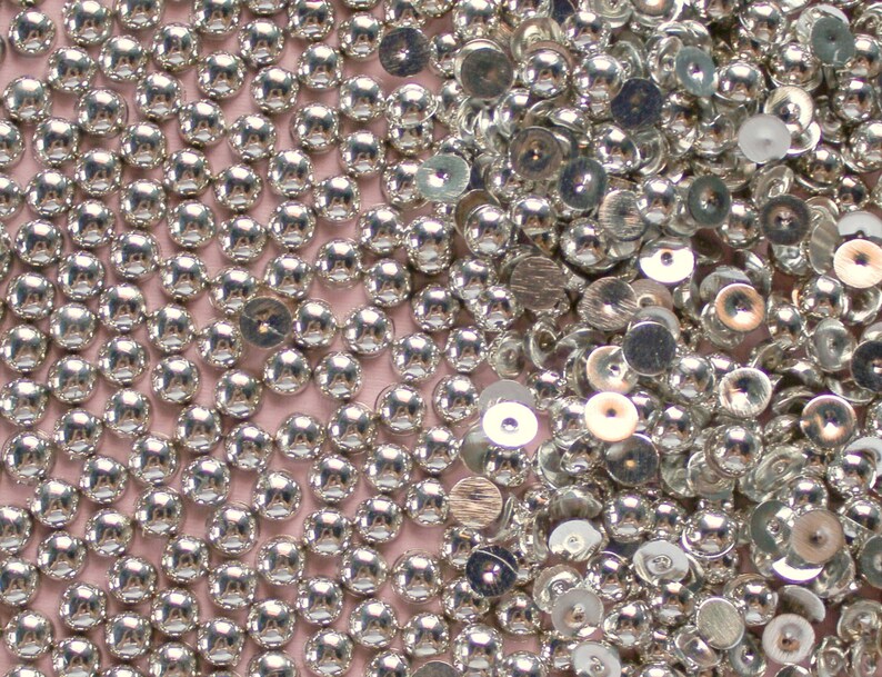 May include: A close-up image of a large quantity of silver-colored round and flat sequins. The sequins are scattered randomly on a pink surface.