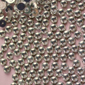 May include: A close-up of a large quantity of silver-colored round metal embellishments. The embellishments are scattered on a light pink surface.