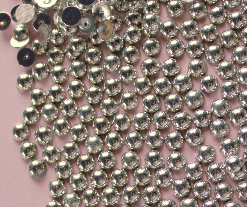 May include: A close-up of a large quantity of silver-colored round metal embellishments. The embellishments are scattered on a light pink surface.