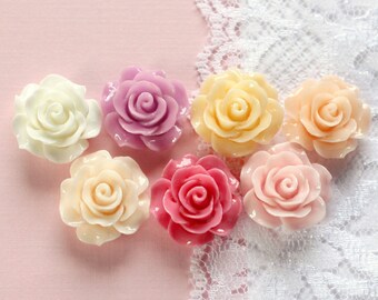 May include: Seven resin rose cabochons in various colors: white, lavender, yellow, peach, and pink. The roses are arranged on a pink surface with a white lace trim. These are ideal for crafting and jewelry making.