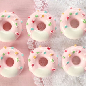 May include: Six pink frosted donuts with white icing and colorful sprinkles. The donuts are arranged in two rows of three on a white and pink background.
