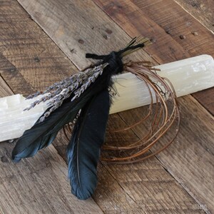 Puede incluir: Una varita de cristal de selenita con un manojo de lavanda seca y plumas negras atadas con un cordel negro.