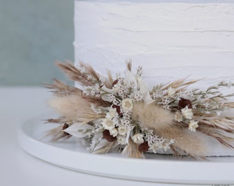 May include: A white cake with a rustic, dried flower arrangement on top. The flowers are a mix of white and brown tones, with some dried grasses and pampas grass.