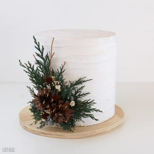 May include: A cylindrical cake with white textured frosting, decorated on one side with a cluster of green cedar sprigs, brown pinecones, and small white dried flowers, all resting on a round light-colored wooden cake board.
