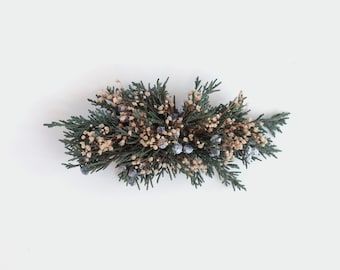 May include: A green and brown floral arrangement with small blue berries. The arrangement is on a white background.