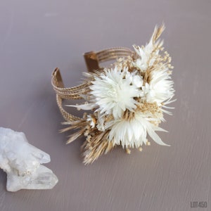 May include: A gold metal bracelet with dried white flowers and tan grasses attached to it. The bracelet is on a gray surface.