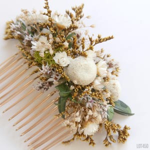 May include: A floral hair comb featuring a variety of dried flowers in shades of white, cream, and brown. The comb has gold-colored metal teeth and includes small green leaves and spherical, textured blooms. The arrangement is dense and detailed.