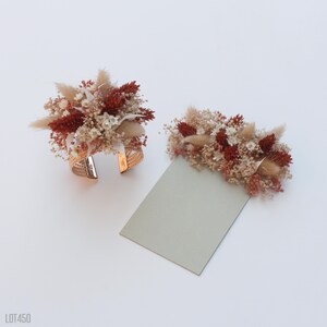 May include: A set of two dried flower arrangements, one on a gold metal cuff bracelet and the other on a light gray card. The flowers are a mix of white, beige, and rust-colored dried flowers.